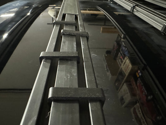 A black plastic cable management keeper installed on a vehicle's roof rack cross bar.