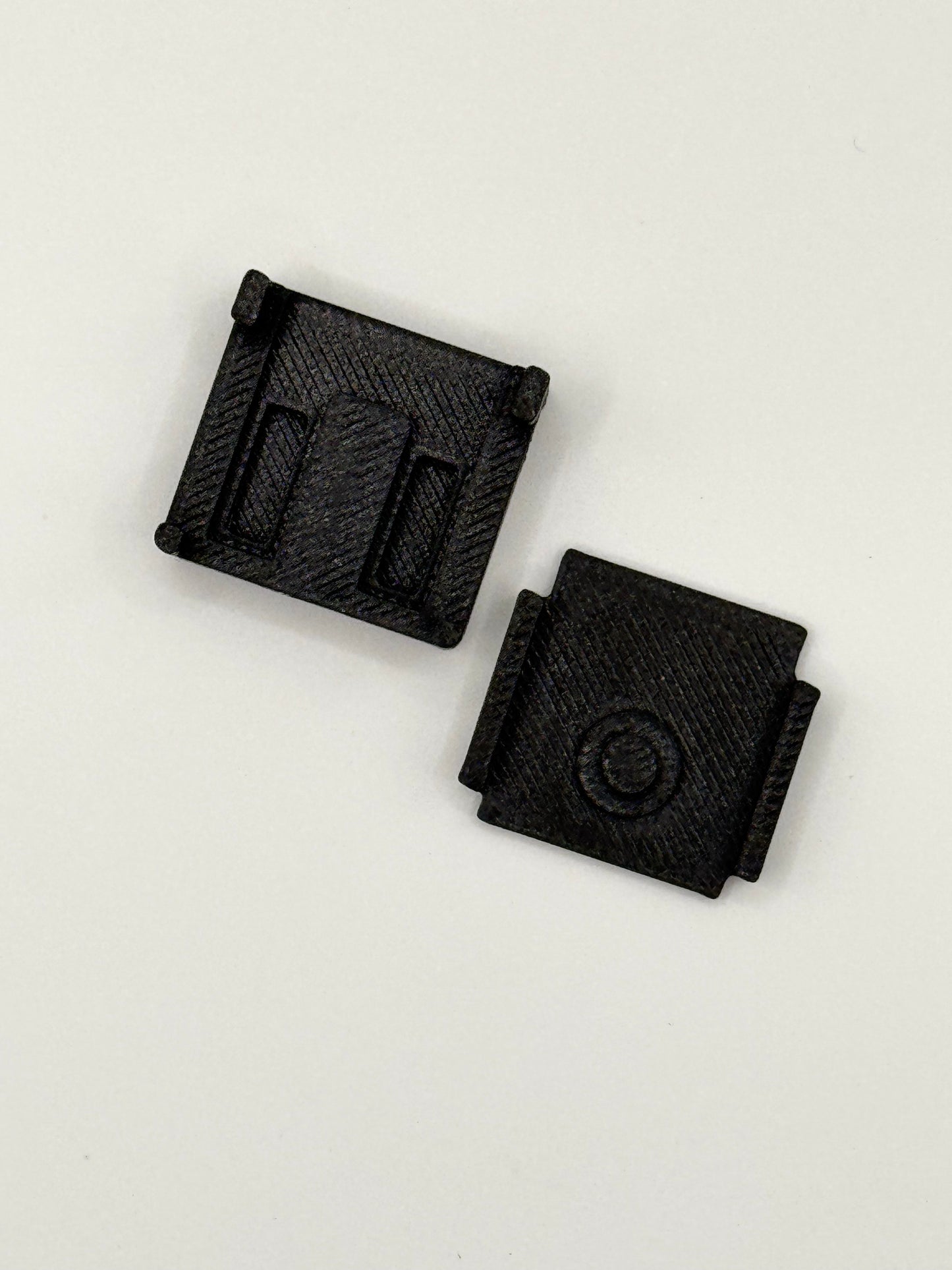 Two small black textured plastic components with rectangular slots and a circular detail, shown on a plain white background.